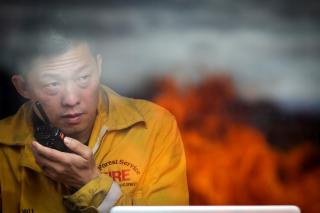 Wildland firefighter, Samuel Wu talking on a radio while fire burns in the background.