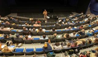 From above/top of the room: Large auditorium with lecturer up front. Participants fill rows of chairs.