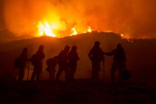 Firefighters stand silohetted as it's night and a fire burns in the forest behind them.