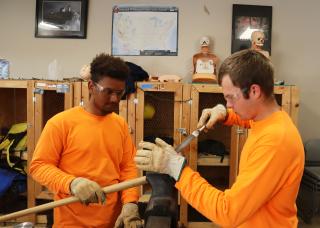 Two young men, wearing long-sleeved orange shirts and safety glasses, work together to sharpen and tools. The one on the left holds the tool while the one on the right sharpens it.