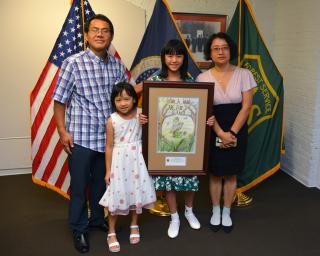 A family (mom, dad, and two girls -one younger than the other) pose for a family portrait. The older girl is holding a frame copy of a Woodsy Owl drawing. Behind them, three flags: US, Forest Service and USDA)