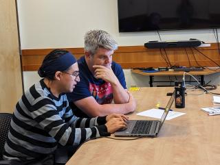 Two men, sittign at a desk inside a conference room, stare together at a laptop.