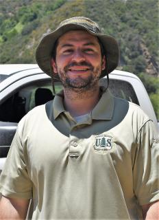 Close-up: Connor Renard, in Forest Service polo and bucket hat, in front of a vehicle with mountains behind him.