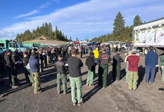 A large group of people gather in a circle with fire trucks to left.