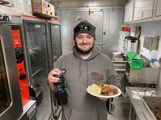 A man, wearing a grey hoodie and ball caps, smiles at the camera as he holds aplate of food on one hand and a bottle of soda on the other