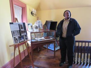 A woman standing inside a historic building, next to an exhibit and some historical artifacts and photos.