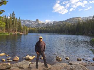 A man poses for the camera by standing on the shores in front of a body of water (lake). Large trees and mountains adorn the far shore behind him (background).