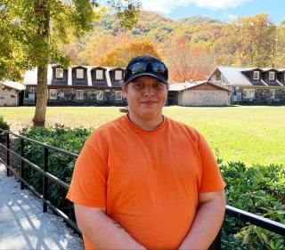 Timothy Hawkins, wearing orange T-shirt and black baseball cap with sunglasses perched on its brim, stands on a sidewalk with Job Corps facilities and tree-covered hill behind him.
