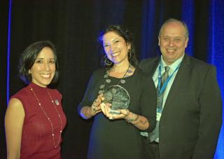 A woman (center) holds an award. A man and another woman stadnd on either sdie of the camera.