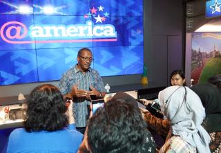 Chief Moore in front of screen displaying @america logo, speaking with Indonesian students.