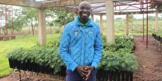 A man stands smiling in front of hundreds of tree seedling in rows of small pots.