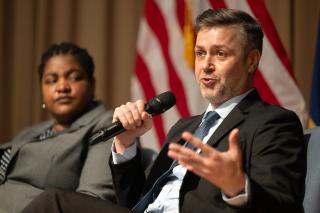 A man sitting down, taking into a microphone from a stage. A woman, sitting next to him, looks on as he speaks.