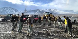 A group of people outside standing in a circle listening to a safety briefing. Snow covered mountains and storm clouds loom in the distance.