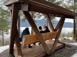 From behind: Three people sit on the bench and enjoy the view of Mendenhall Lake. Snow is still on the ground surrounding the structure.