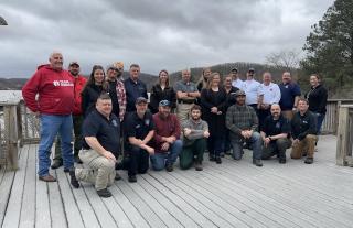 Image shows a group photo of two rows of people outside on a deck, with the front row kneeling down and the second row standing. Everyone is looking at the camera and smiling.