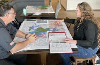 A man and a woman sit on either side of a table with a map spread out between them. The woman is pointing to areas on the map as the man looks on.