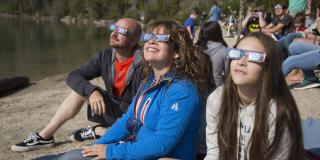 People sit along a sandy lake beach, looking up at at the sun through through safety glasses.
