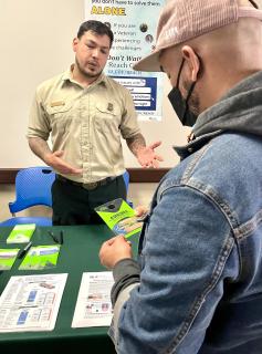 A man in a mask is talking to a man in Forest Service uniform. There is a table with a green table cloth and pamphlets in between them. Them with the mask is holding a pamphlet