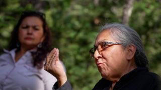 A Native American woman speaks and gestures with her hand. A woman stands listening in the background.