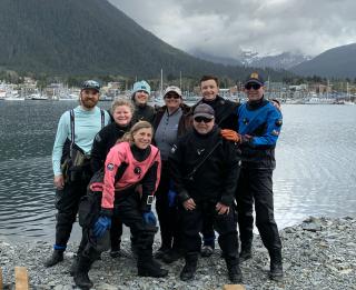 National Diving Control Board participants stand on the edge of water in Sitka, Alaska. A marina is visible on the far shore. In background, Mountains and heavy clouds.