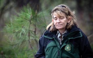 A woman in a forest, wearing a USDA Forest Service jacket, with her eyes closed breathing deeply.