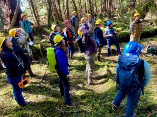 Group of employees in hard hats, carrying backpacks, in forest, looking in the same direction, at an out-of-frame instructor, as part of hands-on training.