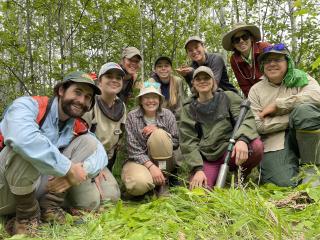 Group photo: People crouched down on grass in forest, looking at camera below them.