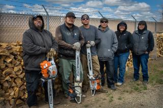 Image shows a group of men and women standing with chainsaws next to a stack of recently cut wood.