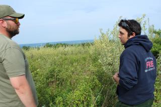 In profile, Forest Service wildlife biologist Greg Flood, wearing sunglasses, t-shirt & baseball cap, speaks to Job Corps student Nate Buck, wearing a hoodie and sunglasses atop his head. They stand at the edge of the field overgrown with honeysuckle.