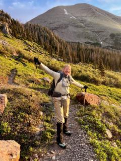 A woman, in FS uniform, standing in the middle of a mountain (peak in the background). Her hands are open as if about to hug and a smaile in her face.