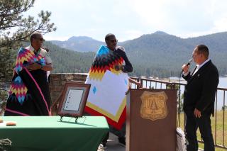 Three men standing up on a podium. One man has a microphone and is addressing the other two, who are wearing colorful native quilts draped over their shoulders.