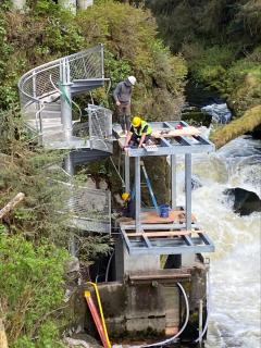 Three workers wearing hard hats assemble the viewing platform while a roaring river rushes below.