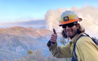 A wildland firefighter/EMT, smiling, holding a thumbs up while a wildfire burns on a distant hilltop.