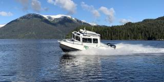A medium sized patrol boat with enclosed cab moving swiftly across a body of water. A snow capped mountain and evergreen forest loom in the background.