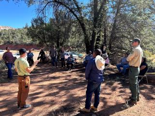A group of people gathers in a large circle on dirt land with trees in the background.
