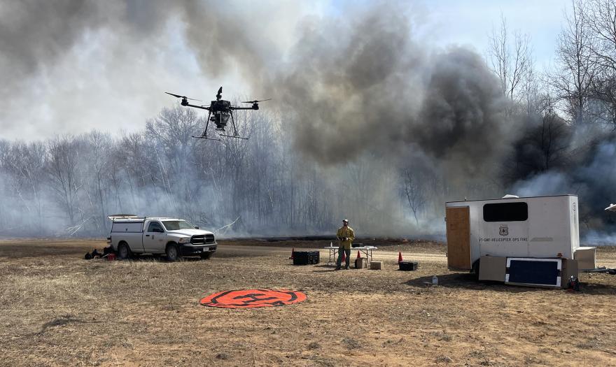 A drone flying in the middle of the photo. Behind the drone,  vehicles with equipment and in the background , black and grey smoke flowing from somewhere off-frame.