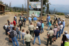 A group of possibly 30 people standing around in a circle in front of a large sign and map on top of a mountain area.