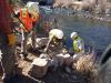 A team of people wearing hard hats, high visibility jackets, and gloves work to move stone blocks on the bank of a river.