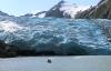 A small boat sailing back from a wall of ice along a lakeshore with mountains in the background.