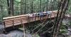 A newly built bridge on a forested trail with the people who built the bridge standing on it and smiling at the camera.