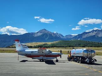 An airplane sits on the tarmac next to a fuel truck with mountains in the background.
