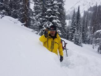 A man is waist deep in snow with pine trees behind him