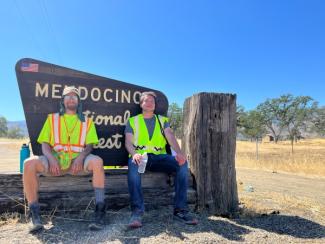 Two people in bright yellow vests rest in shade of Mendocino National Forest sign