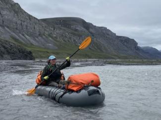 A person paddles a pack raft along the shore with mountains behind.