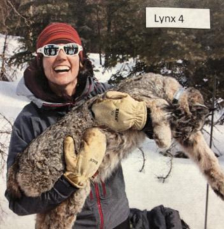a woman wearing a winter coat and thick mittens cradles a lynx in her arms. She is standing in a snowy Forest.