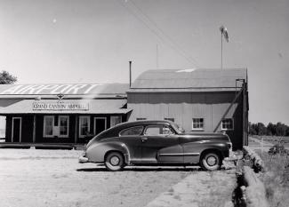 A black-and-white picture of an old car parked in front of a metal airport terminal building.