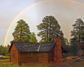 A log cabin sits in front of trees with a rainbow in the sky behind.