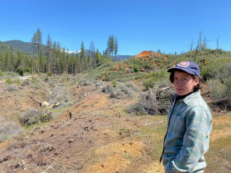 A woman stands to in the left of a photo looking towards the camera with a background of a gully with brush piled for burning and conifer trees in the background.