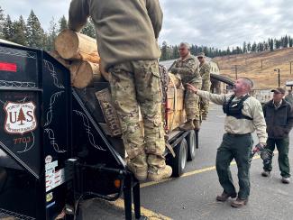 Soldiers and Forest Service employees unload timber logs from the back of a flatbed trailer.