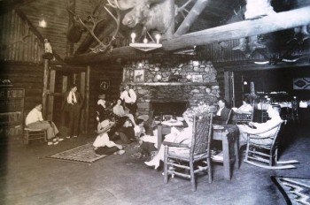 A black and white photo of 14 people in a large room in a lodge by a fireplace. Four people are sitting in rocking chairs.